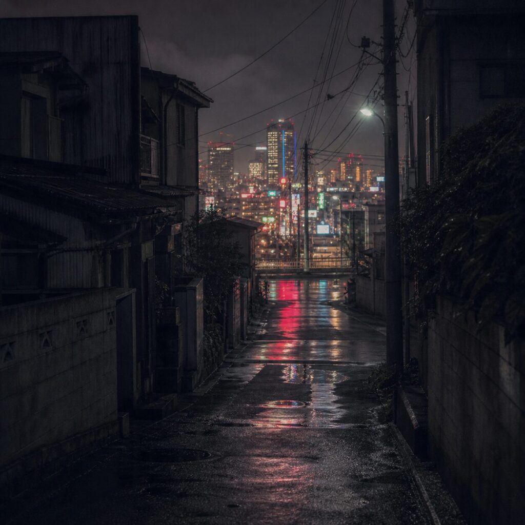暗い路地と濡れた道路の向こうに、1980年代の日本の街を思わせるネオンやビルの明かりが遠くぼんやり輝いている画像